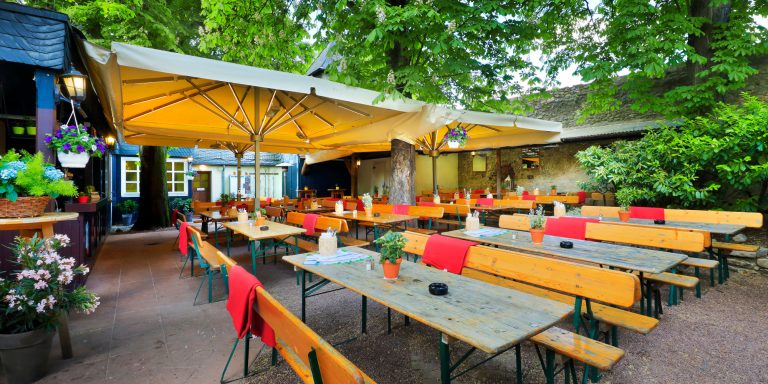 Blick auf einen Garten mit langen Holztischen und roten Tischdecken unter Sonnenschirmen.